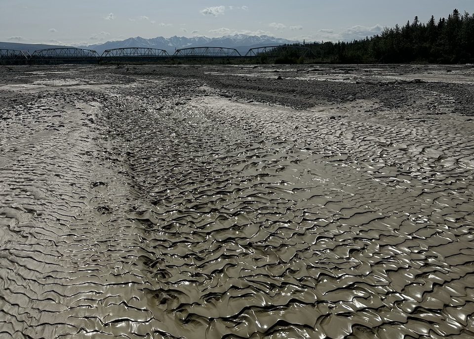Silt-covered, tan ripples of the Gertzle River, Alaska, in the foreground, with a bridge and the Alaska Range mountains in the distant background