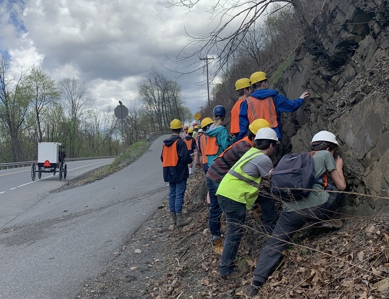 Group of geology students in bright, high-visibility vests and helmets observing a Pennsylvania roadside outcrop with a horse-driven buggy driving past in the background