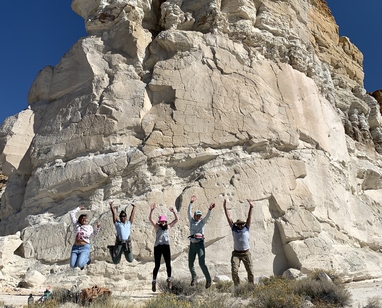 Five geologists leaping joyfully in front of a white-tan rock outcropping under a clear blue sky in the Wind River Basin, Wyoming