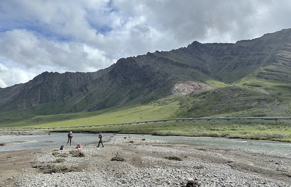 Two geologists measuring sediments on the braided Atigun River, Alaska; dark, layered mountains in the background grade into green, sloping tundra; Alaska oil pipeline visible between the river and the mountains